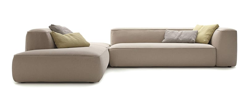 En modern L-formad beige sektionssoffa, som påminner om snygg design som finns i en presentbutik i Göteborg, har tre kuddar i grått och gult. Mot en vit bakgrund har den rena linjer och en minimalistisk estetik.