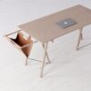 Ett minimalistiskt skrivbord i trä med en beige laptop sitter elegant och liknar något du kan hitta i en chic Göteborgs presentbutik. Dess unika integrerade hängmatta-liknande läderhylla på ena sidan, stödd av trästänger, kompletterar golvets ljusa träton.