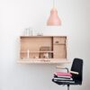 En minimalistisk arbetsyta med ett väggmonterat skrivbord i trä med hyllor med böcker och pappersvaror under en rosa taklampa. En svart stol, accentuerad med en souvenir från Haga, sitter framför skrivbordet, sin sits prydd med en hög böcker.
