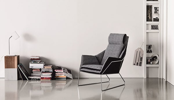 Ett modernt, minimalistiskt rum med en grå fåtölj och en elegant golvlampa. Ett litet sidobord visar en souvenir från Göteborgs presentbutik, medan en smal vägghylla rymmer böcker och inredning. Det polerade golvet återspeglar den fridfulla elegansen i detta eleganta utrymme.