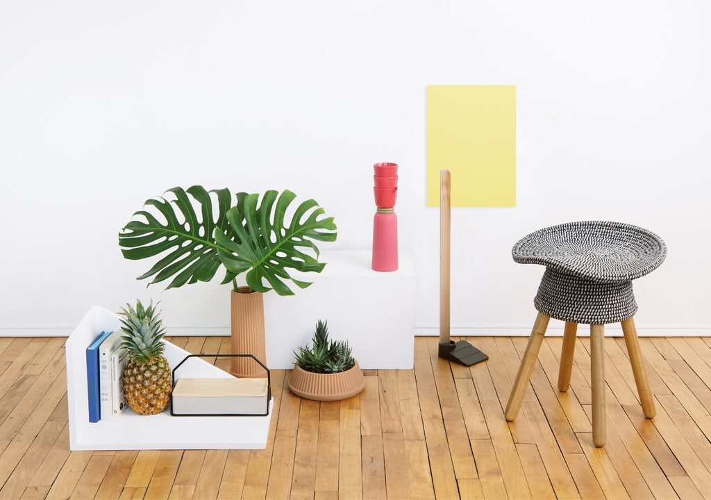 En modern, minimalistisk uppsättning med flätad pall, krukväxter och dekorativa föremål som påminner om en presentbutik i Göteborg. Ananas och böcker vilar på trägolvet, medan en vit bakgrund och gult papper förgyller scenen.