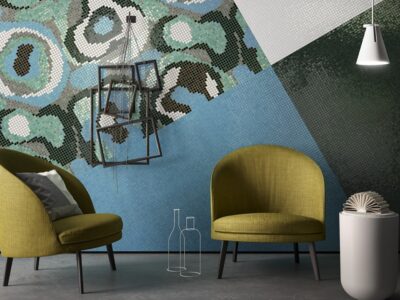 Ett modernt rum med två gröna stolar, ett litet vitt bord med en bok och en modern lampa. Väggen har ett färgstarkt, abstrakt geometriskt mönster i blått, grönt och grått, med en unik fyrkantig ram kanske hämtad från en Göteborgs presentbutik.