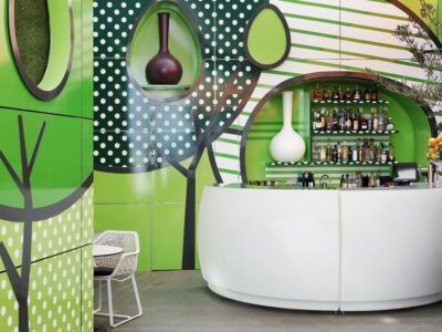 En modern bar i Göteborg, med en vit, svängd disk, omgiven av dekorativa gröna paneler med abstrakt träddesign. På hyllorna bakom disken finns olika flaskor och två vaser. I förgrunden erbjuder ett litet bord och en stol en mysig plats att njuta av din souvenirdrink.