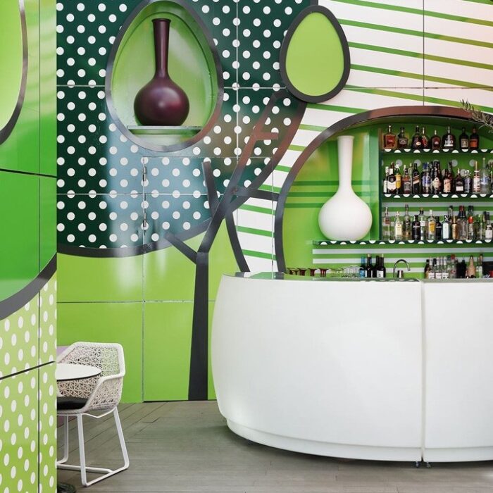 En modern bar i Göteborg, med en vit, svängd disk, omgiven av dekorativa gröna paneler med abstrakt träddesign. På hyllorna bakom disken finns olika flaskor och två vaser. I förgrunden erbjuder ett litet bord och en stol en mysig plats att njuta av din souvenirdrink.