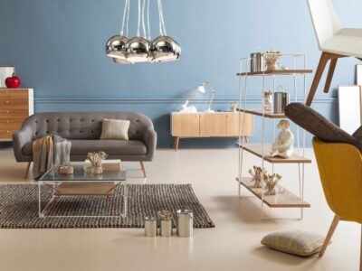 Ett modernt vardagsrum har en grå soffa, ett soffbord i metall och glas, tvåfärgade hyllor prydda med Göteborgssouvenirer och en gul stol. Snygga taklampor hänger i taket. De blå väggarna och det ljusa golvet skapar en mysig atmosfär.
