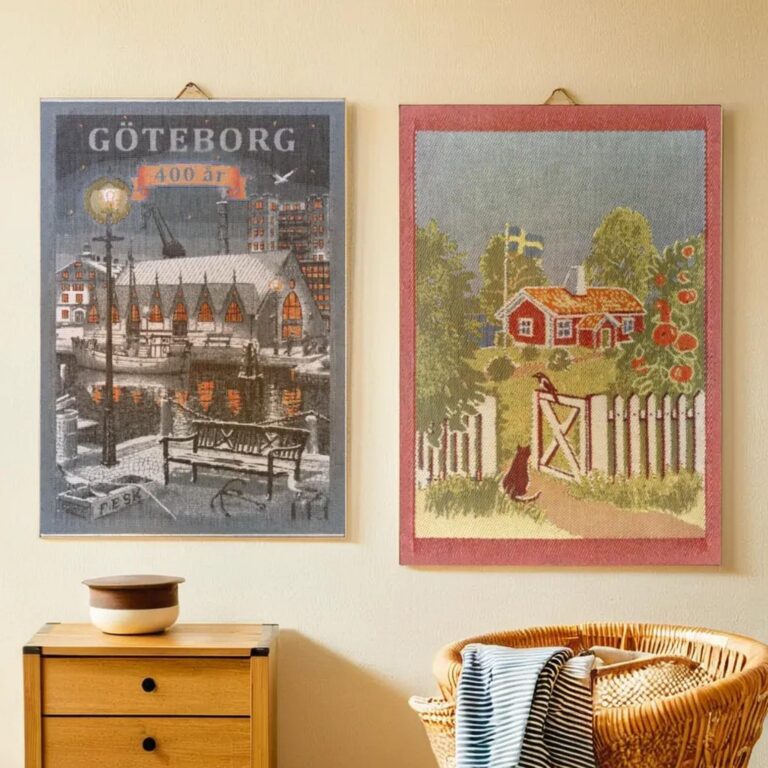 Två inramade konstverk hänger på en beige vägg ovanför ett träskåp och en flätad korg. Det vänstra konstverket föreställer en nattlig stadsbild med texten "Göteborg 400 år." Det högra konstverket skildrar en landsbygdsscen med en stuga, träd och blommor.