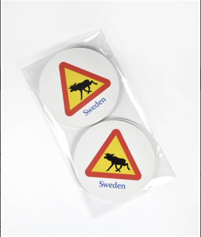 Två cirkulära underlägg med en gul och röd triangulär varningsskylt för älg och ordet "Sverige" tryckt nedanför, förpackade tillsammans i ett genomskinligt plastfodral mot vit bakgrund.