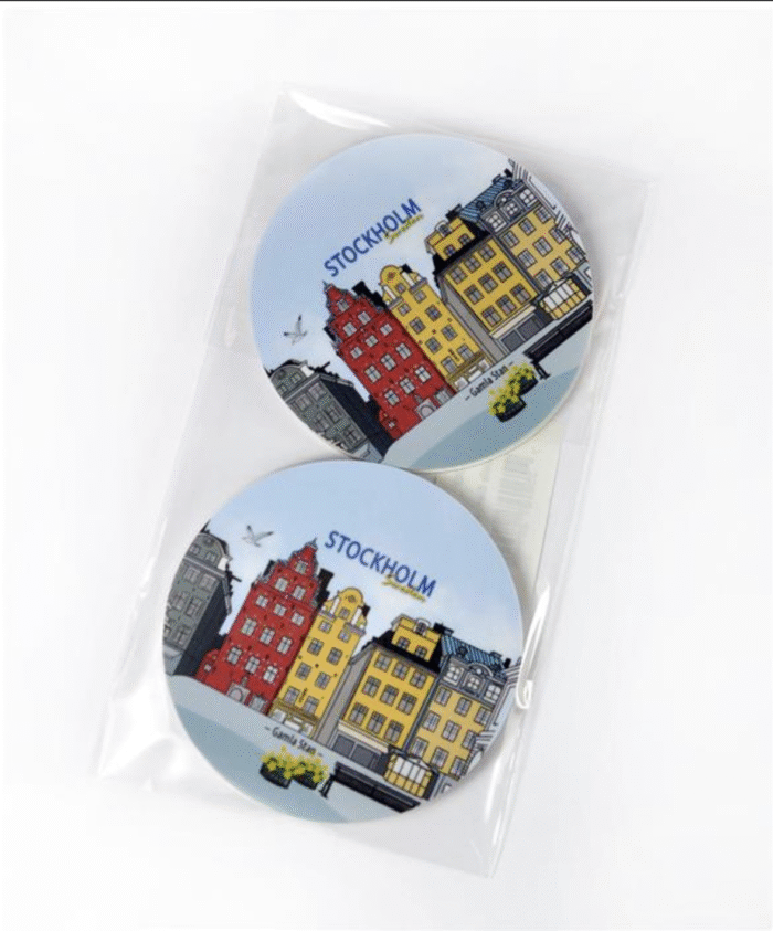 Två runda underlägg med färgglada byggnader i Stockholms stadskärna och texten ”STOCKHOLM” ovanför, placerade i ett genomskinligt plastfodral mot vit bakgrund.