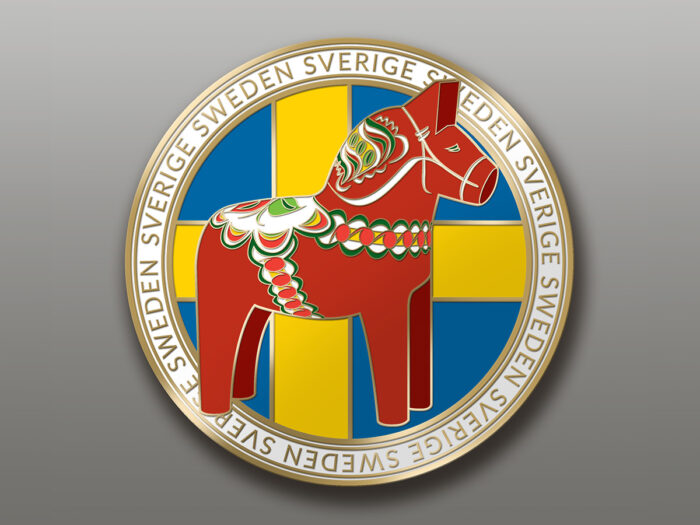 Ett runt emblem föreställer en röd dalahäst med mönster på en gul och blå svensk flaggbakgrund, kantad av ”SWEDEN SVERIGE” i guld på vitt. Produkt: Husky, liggande 25 cm.